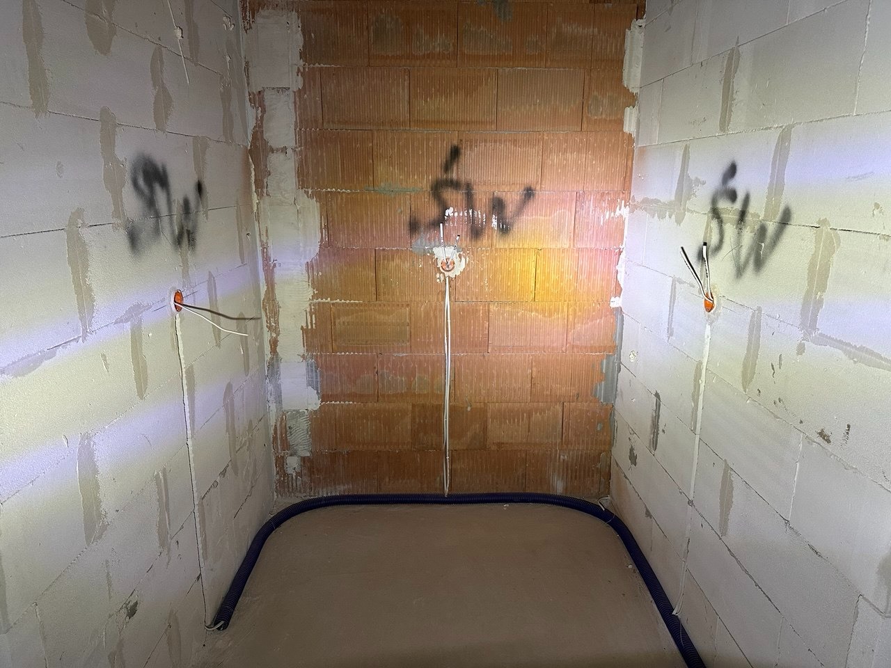 Surowy stan łazienki z instalacją elektryczną w ścianach z betonu komórkowego i rurą karbowaną w podłodze. Widoczne oznaczenia na ścianach.