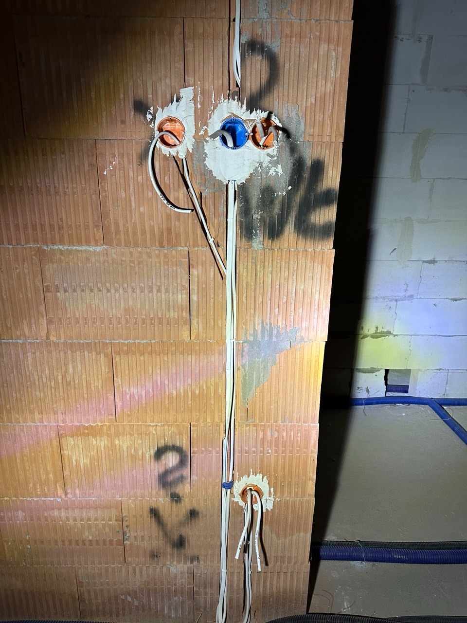 Instalacja elektryczna w trakcie montażu: przewody w peszlach wychodzące ze ściany z pustaków ceramicznych, puszki instalacyjne wmurowane w ścianę, widoczne oznaczenia na murze.