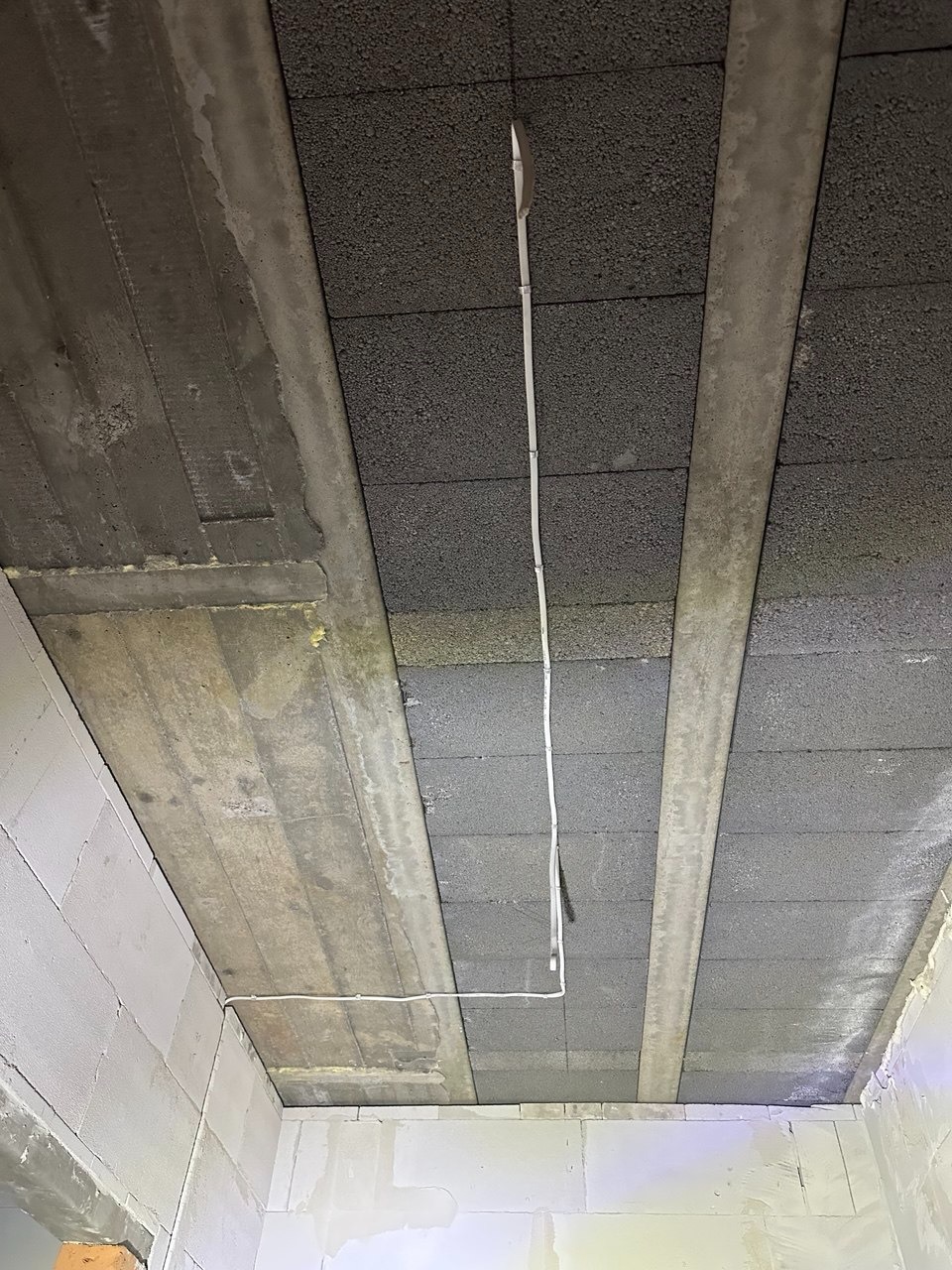 Surowy strop z betonowych belek i pustaków, z poprowadzoną białą instalacją elektryczną. Widoczne ściany z białych bloczków. Ujęcie z dołu, w trakcie prac wykończeniowych.
