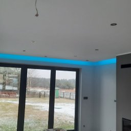 Usługi budowlane - Nowoczesne wnętrze z podwieszanym sufitem i oświetleniem LED w kolorze niebieskim, widok na okno z ogrodem w tle. Minimalistyczny design.