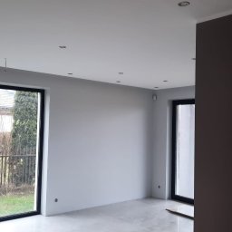 Usługi budowlane - Jasne, minimalistyczne wnętrze po remoncie z dużymi oknami i szarą kolorystyką ścian. Widoczne nowe panele podłogowe i oświetlenie sufitowe.