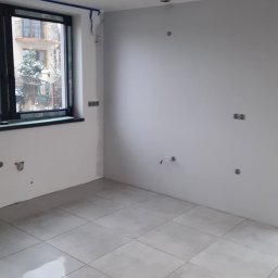 Usługi budowlane - Jasne, kwadratowe płytki podłogowe w pokoju z oknem. Ściany pomalowane na biało, widoczne punkty oświetleniowe w suficie i wystające kable. Okno z niebieską taśmą malarską.