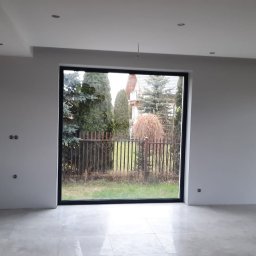 Usługi budowlane - Wnętrze po remoncie w Stroniu Śląskim: jasne ściany, szara podłoga z płytek, duże okno z widokiem na ogród. Widoczne gniazdka elektryczne i punkty świetlne.