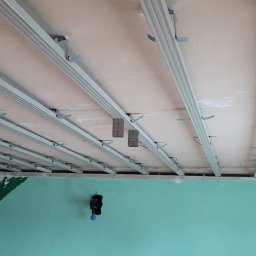 Usługi budowlane - Stelaż sufitu podwieszanego z płyt karton-gips, widoczne profile i uchwyty montażowe, ściany w kolorze miętowym z dekoracyjnymi elementami.