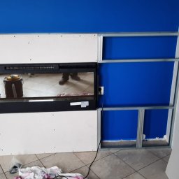 Usługi budowlane - Montaż wkładu kominkowego z obudową z płyt gipsowo-kartonowych na metalowym stelażu, widoczne niebieskie ściany i fragment podłogi z płytek.