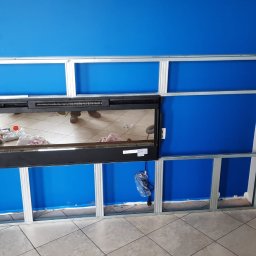 Usługi budowlane - Metalowa konstrukcja pod zabudowę kominka na tle niebieskiej ściany. Widoczne profile i fragment kominka. Prace wykończeniowe w trakcie realizacji.