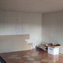 Usługi budowlane - Wnętrze w trakcie prac: ściany obłożone płytami karton-gips, widoczne łączenia i otwory na instalacje. Na podłodze leżą płyty i narzędzia.