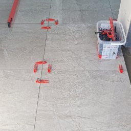 Usługi budowlane - Układanie płytek podłogowych z użyciem systemu poziomowania. Widoczna poziomica, wiadro z narzędziami i fragment stopy osoby wykonującej prace.