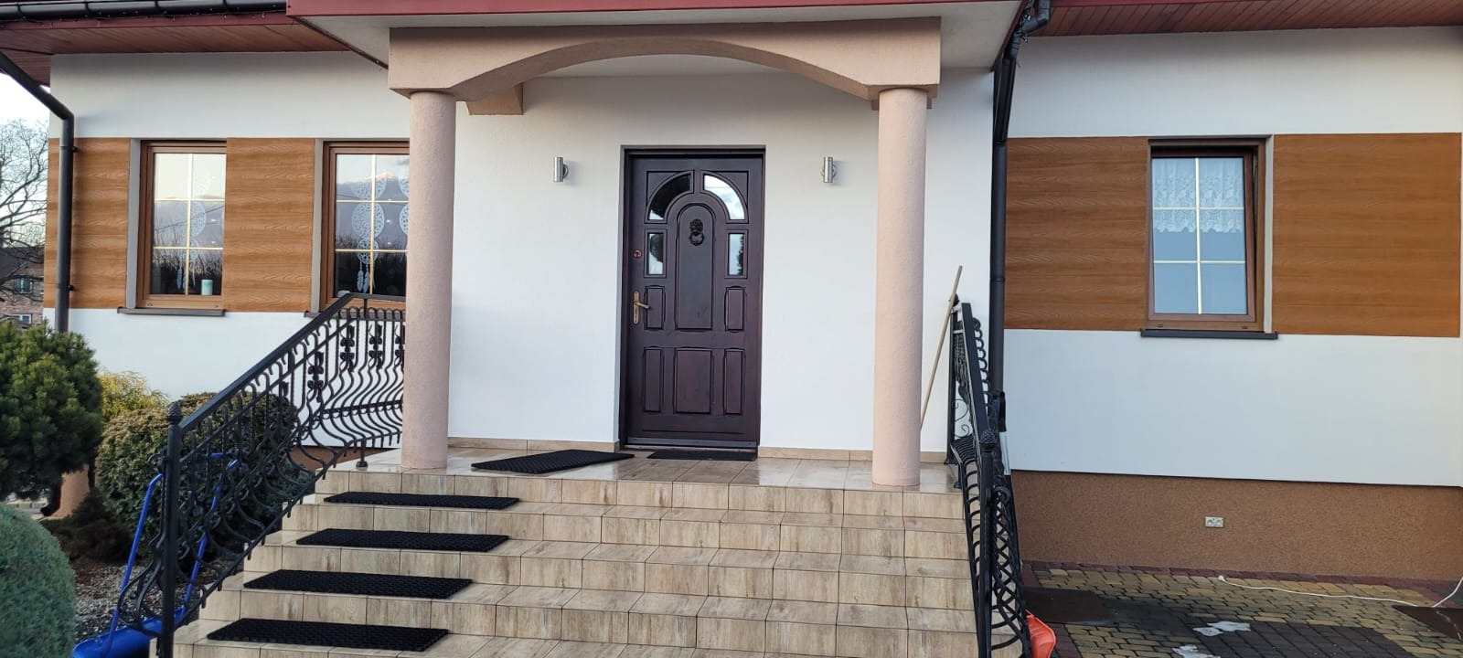 Elewacja domu jednorodzinnego z brązowymi panelami, schodami wejściowymi i ciemnymi drzwiami. Widoczne okna z drewnianą ramą i kolumny podtrzymujące zadaszenie.