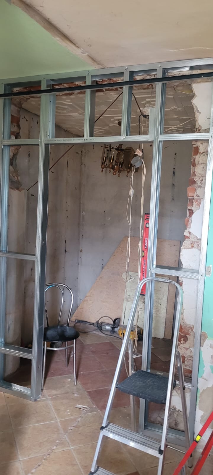 Metalowa konstrukcja szkieletowa ścianki działowej w trakcie montażu, widoczne narzędzia i elementy wyposażenia wnętrza w surowym stanie. Fragment starej instalacji elektrycznej.