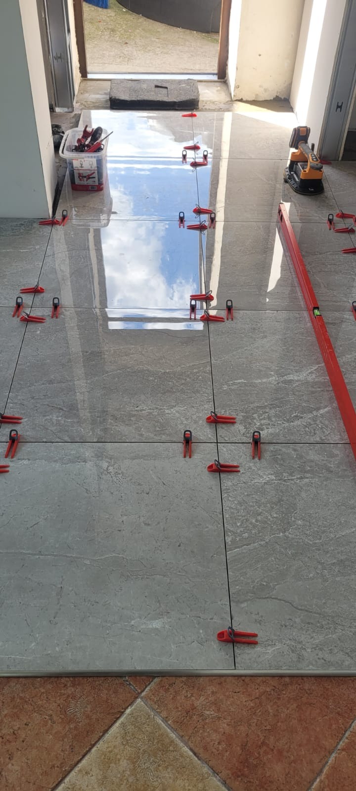 Ułożone szare płytki podłogowe z czerwonymi klipsami poziomującymi, widoczna poziomica i narzędzia glazurnicze. W tle widok na zewnątrz.