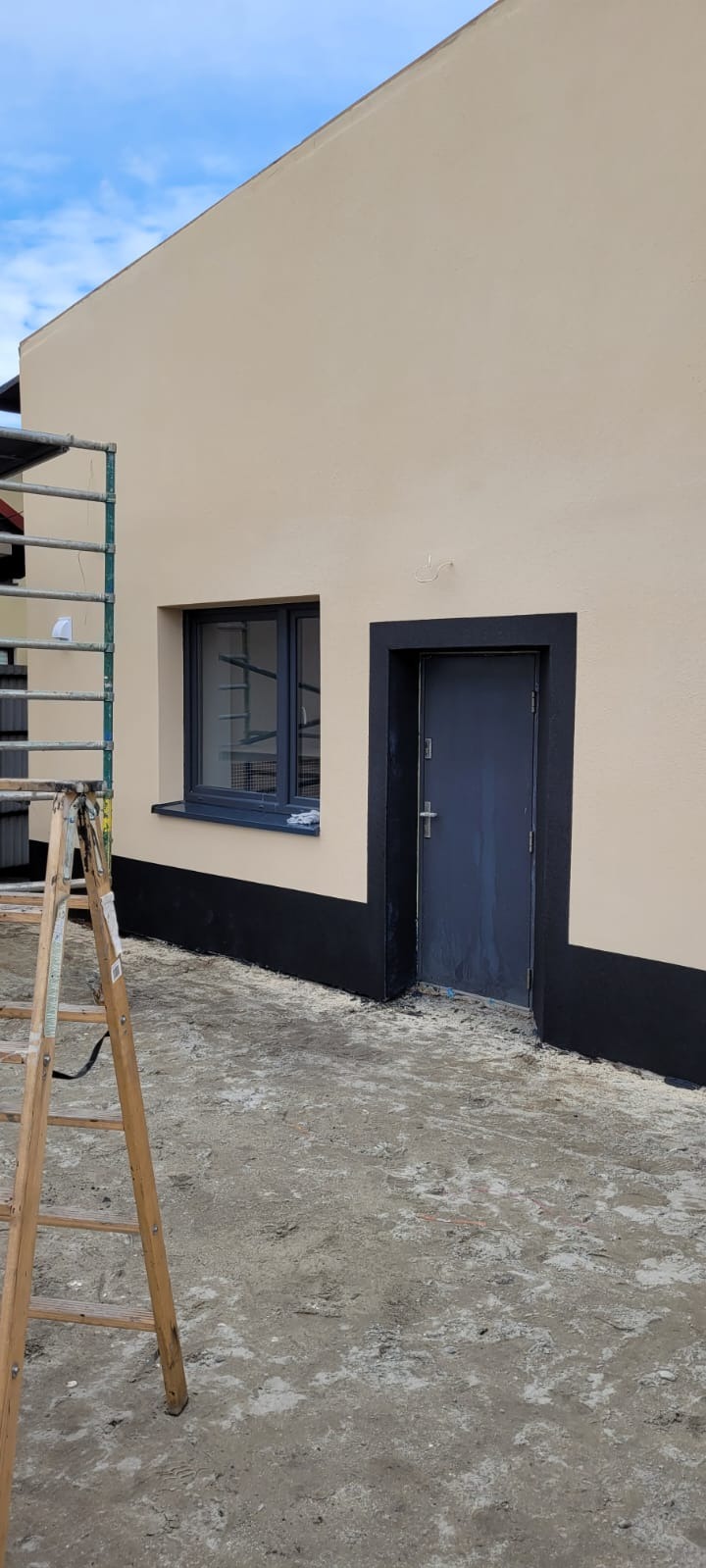 Elewacja budynku po malowaniu z czarnym cokołem i obramowaniem okna oraz drzwi. Drewniana drabina oparta o ścianę. Widok na fragment budynku w słoneczny dzień.