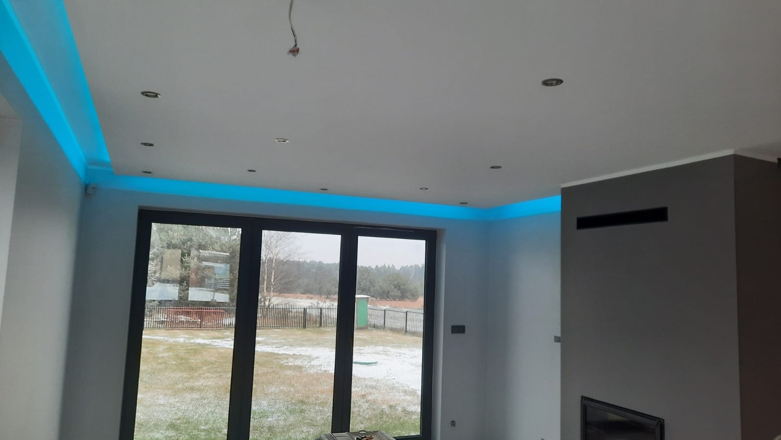 Nowoczesne wnętrze z podwieszanym sufitem i oświetleniem LED w kolorze niebieskim, widok na okno z ogrodem w tle. Minimalistyczny design.