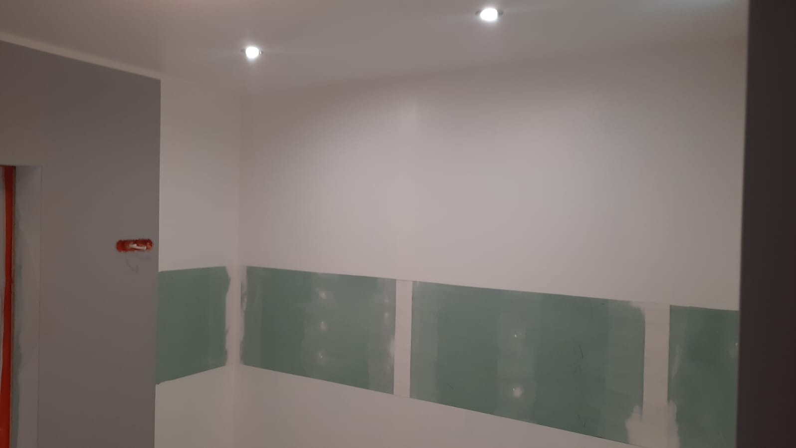 Ściana z zamontowanymi płytami kartonowo-gipsowymi, przygotowana do dalszych prac wykończeniowych. Widoczne punkty oświetleniowe w suficie.