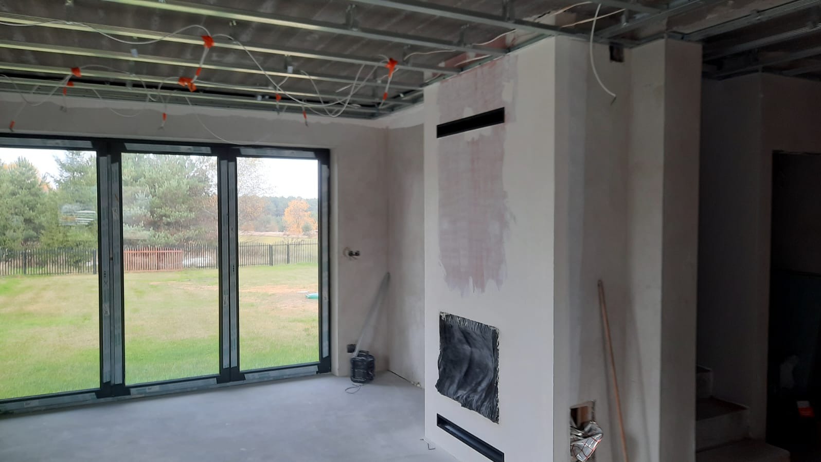 Wnętrze w trakcie wykańczania: sufit podwieszany, instalacja elektryczna, ściany przygotowane do malowania, duże okno z widokiem na zewnątrz. Widoczny kominek w trakcie budowy.