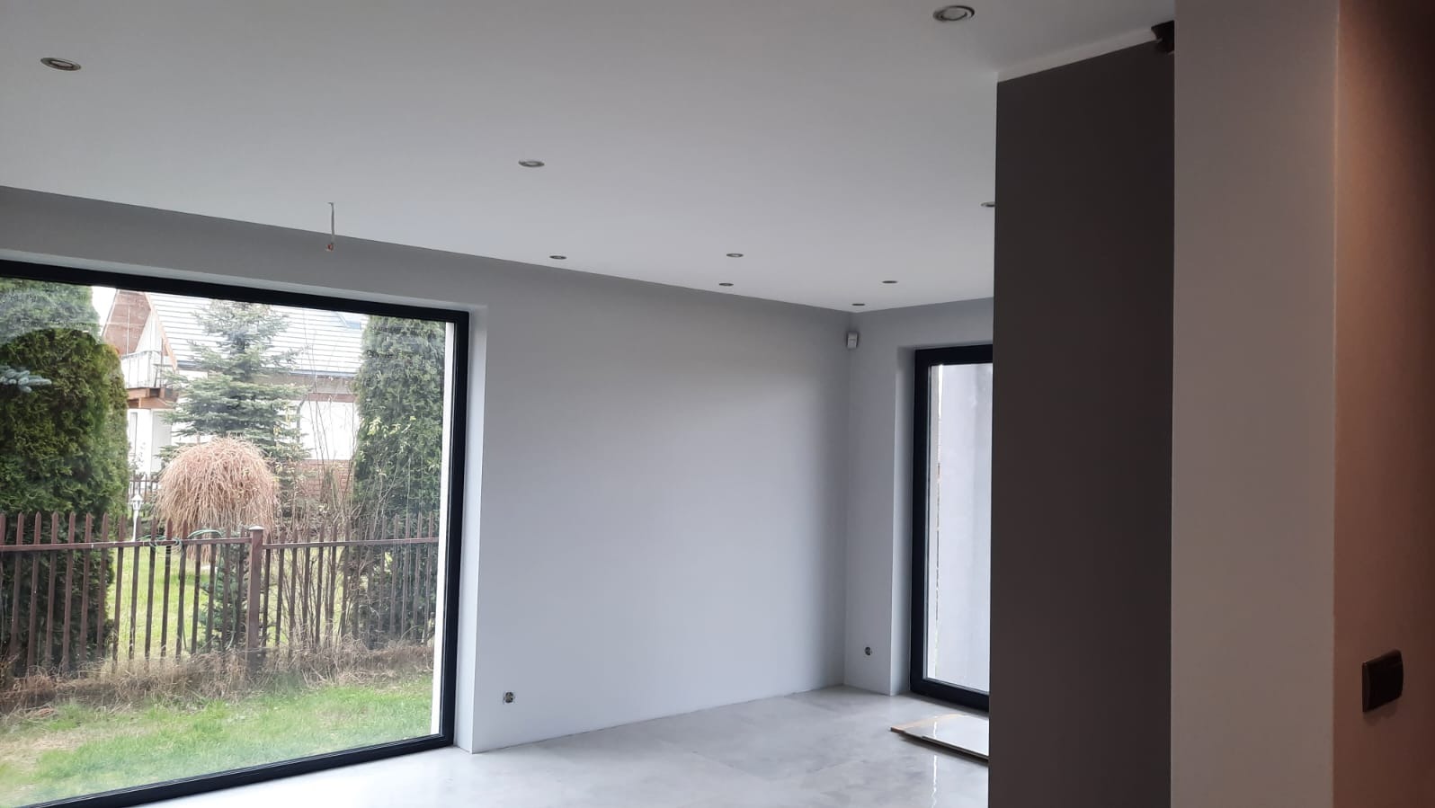 Jasne, minimalistyczne wnętrze po remoncie z dużymi oknami i szarą kolorystyką ścian. Widoczne nowe panele podłogowe i oświetlenie sufitowe.