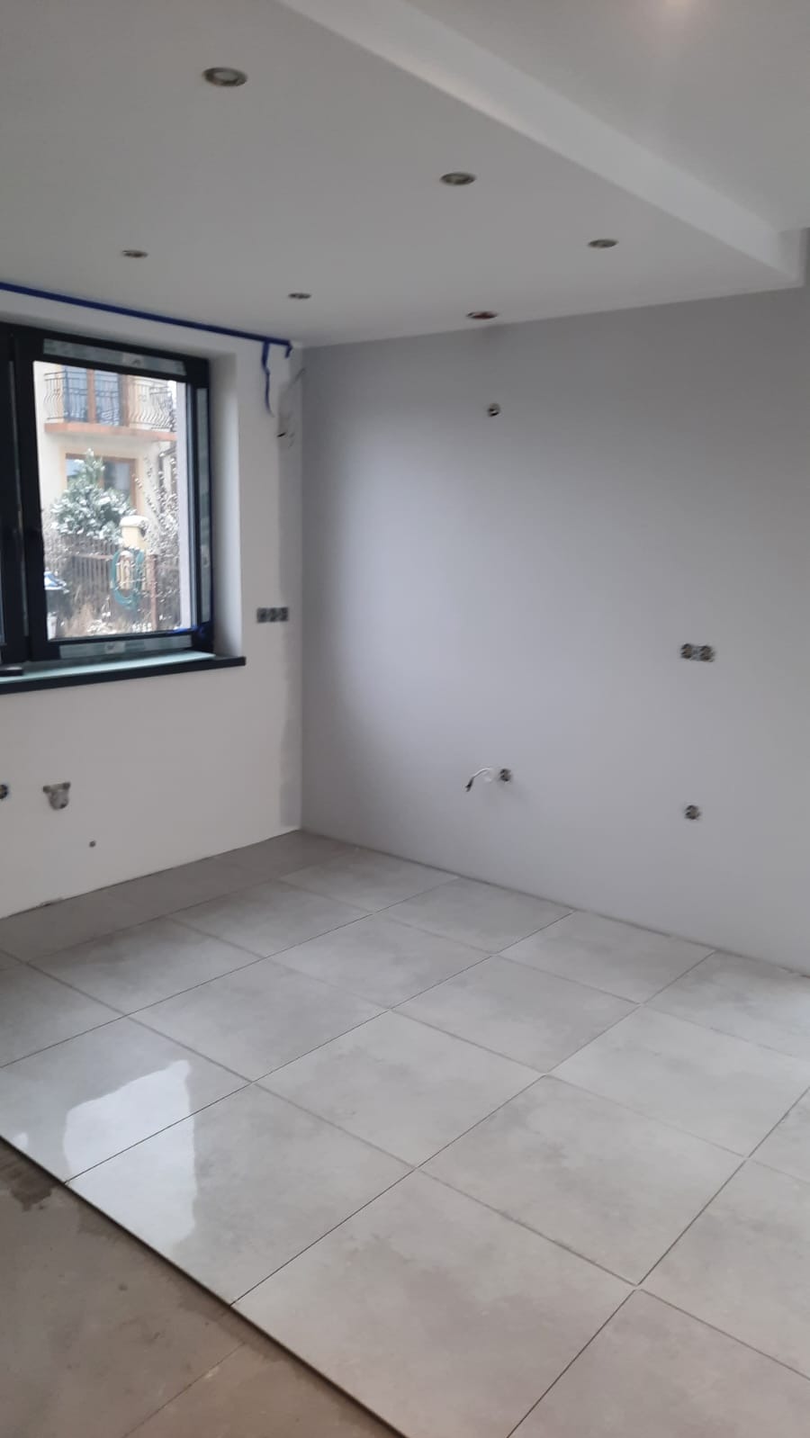 Jasne, kwadratowe płytki podłogowe w pokoju z oknem. Ściany pomalowane na biało, widoczne punkty oświetleniowe w suficie i wystające kable. Okno z niebieską taśmą malarską.