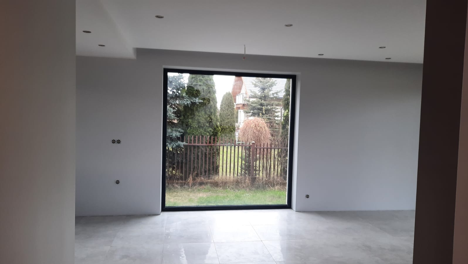 Wnętrze po remoncie w Stroniu Śląskim: jasne ściany, szara podłoga z płytek, duże okno z widokiem na ogród. Widoczne gniazdka elektryczne i punkty świetlne.