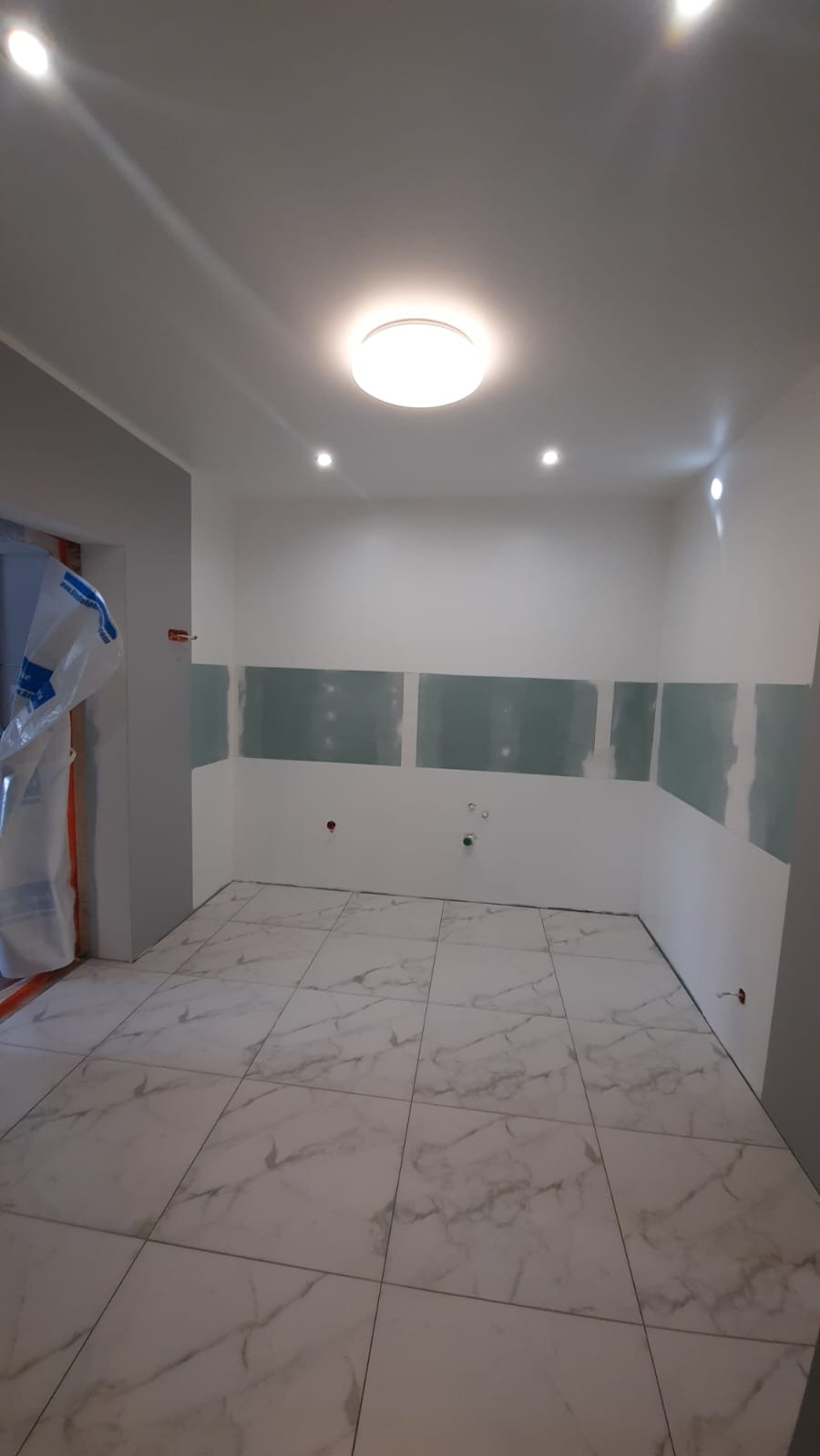 Wnętrze w trakcie prac: białe płytki podłogowe z marmurkowym wzorem, fragmenty płyt karton-gips na ścianach, widoczne instalacje. Oświetlenie sufitowe.