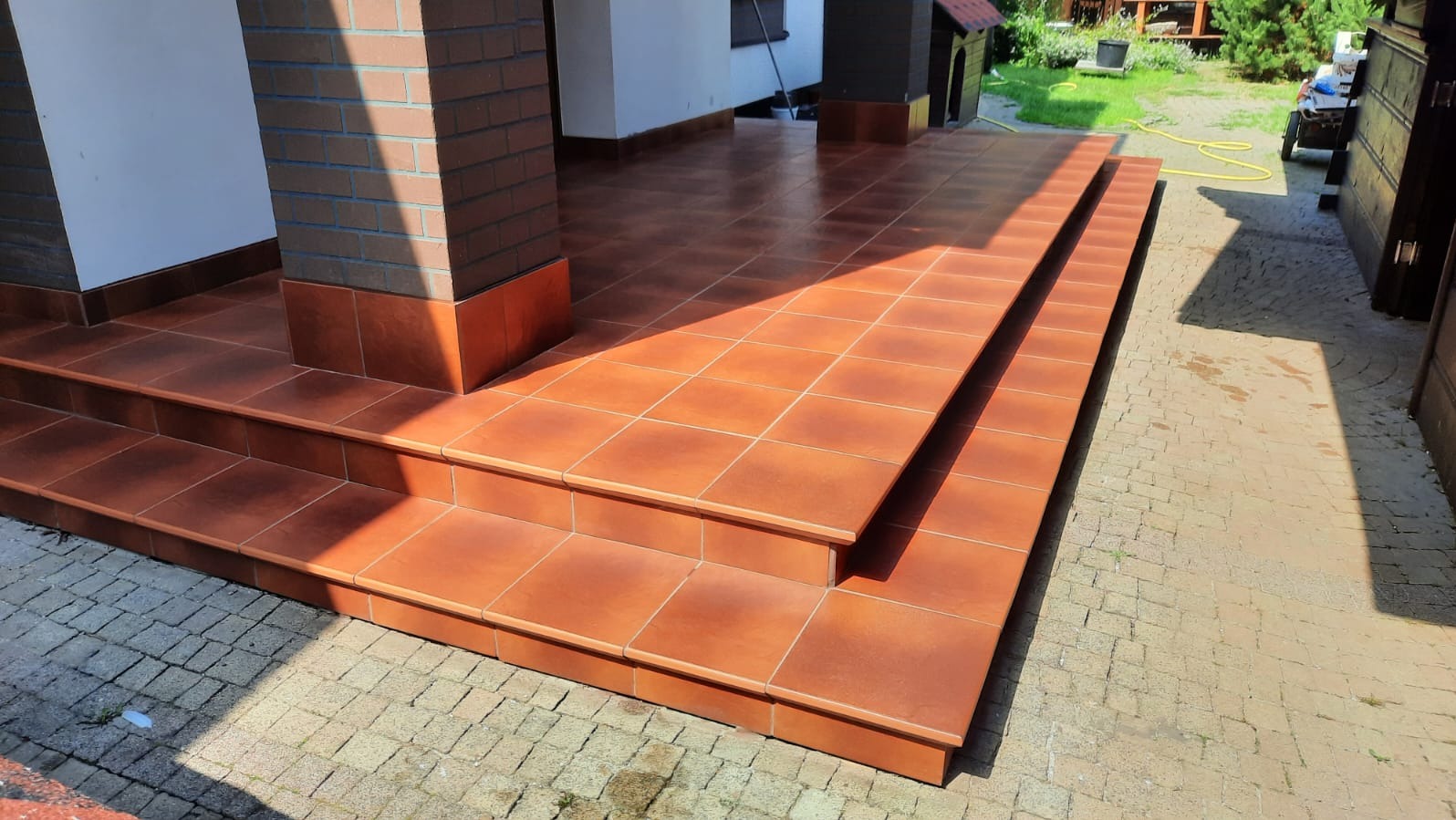 Wykończony taras z brązowych płytek, schody wejściowe. Widoczne kolumny z cegły i fragment ogrodu w tle. Ujęcie z niskiej perspektywy.