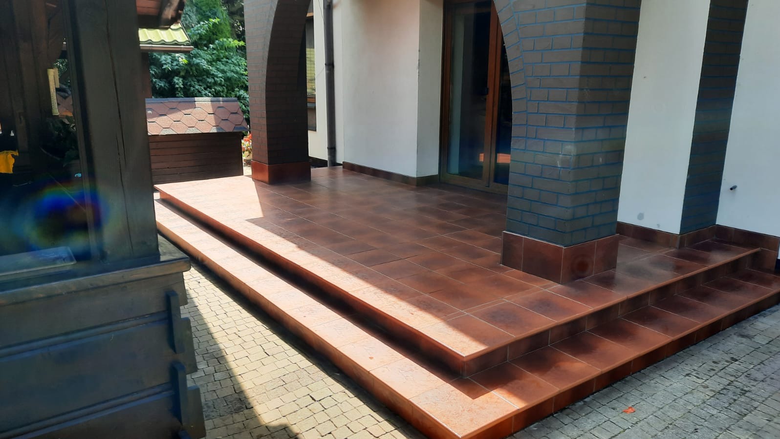 Wykończony taras z brązowych płytek, schody wejściowe z płytek, kolumny z cegły, jasna elewacja budynku. Widok z dołu, w tle zieleń.