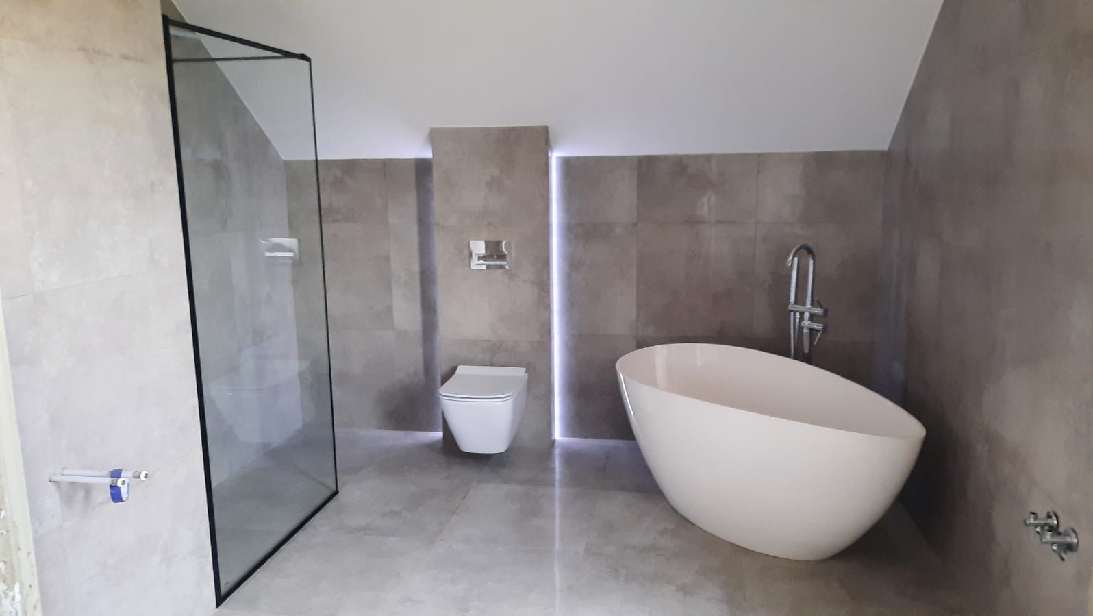 Nowoczesna łazienka z wolnostojącą wanną, szklaną kabiną prysznicową i podświetleniem LED. Minimalistyczny design z szarymi płytkami i białymi elementami wyposażenia.