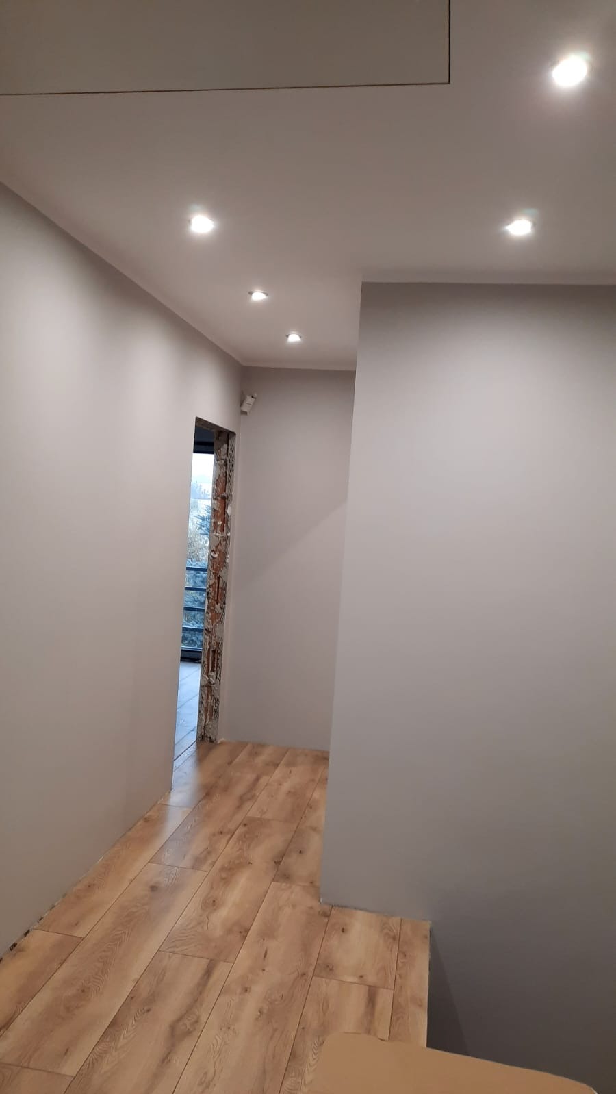 Wykończony korytarz z panelami podłogowymi w odcieniach brązu i beżu. Ściany pomalowane na szaro. Widoczne oświetlenie sufitowe i fragment przejścia z odsłoniętą cegłą.