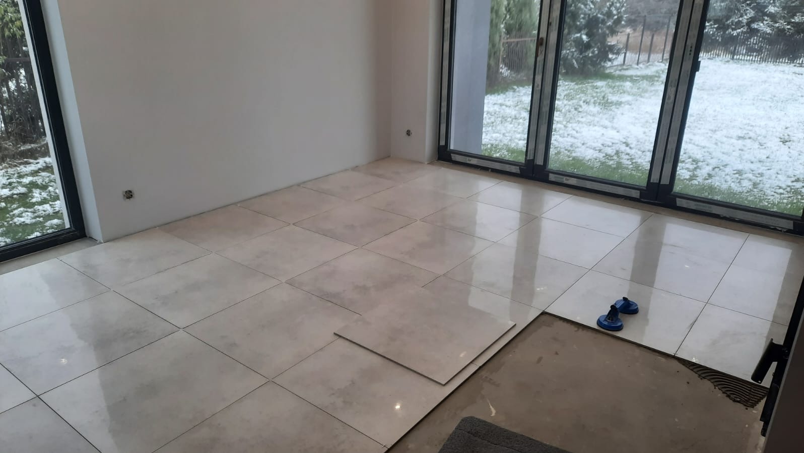 Jasne, kwadratowe płytki podłogowe w trakcie układania w przestronnym pokoju z dużymi oknami. Widoczne przyssawki do płytek i fragment nieukończonej podłogi.