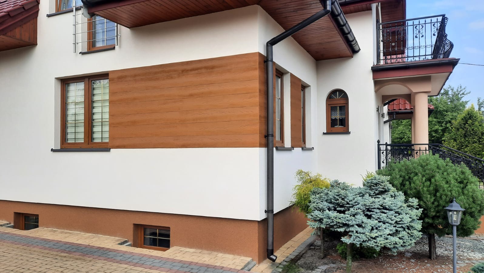 Elewacja domu z drewnopodobną okładziną, białymi ścianami i brązowym cokołem. Widoczne okna z brązowymi ramami i czarna rynna. Uwagę przykuwa starannie wykonana elewacja.