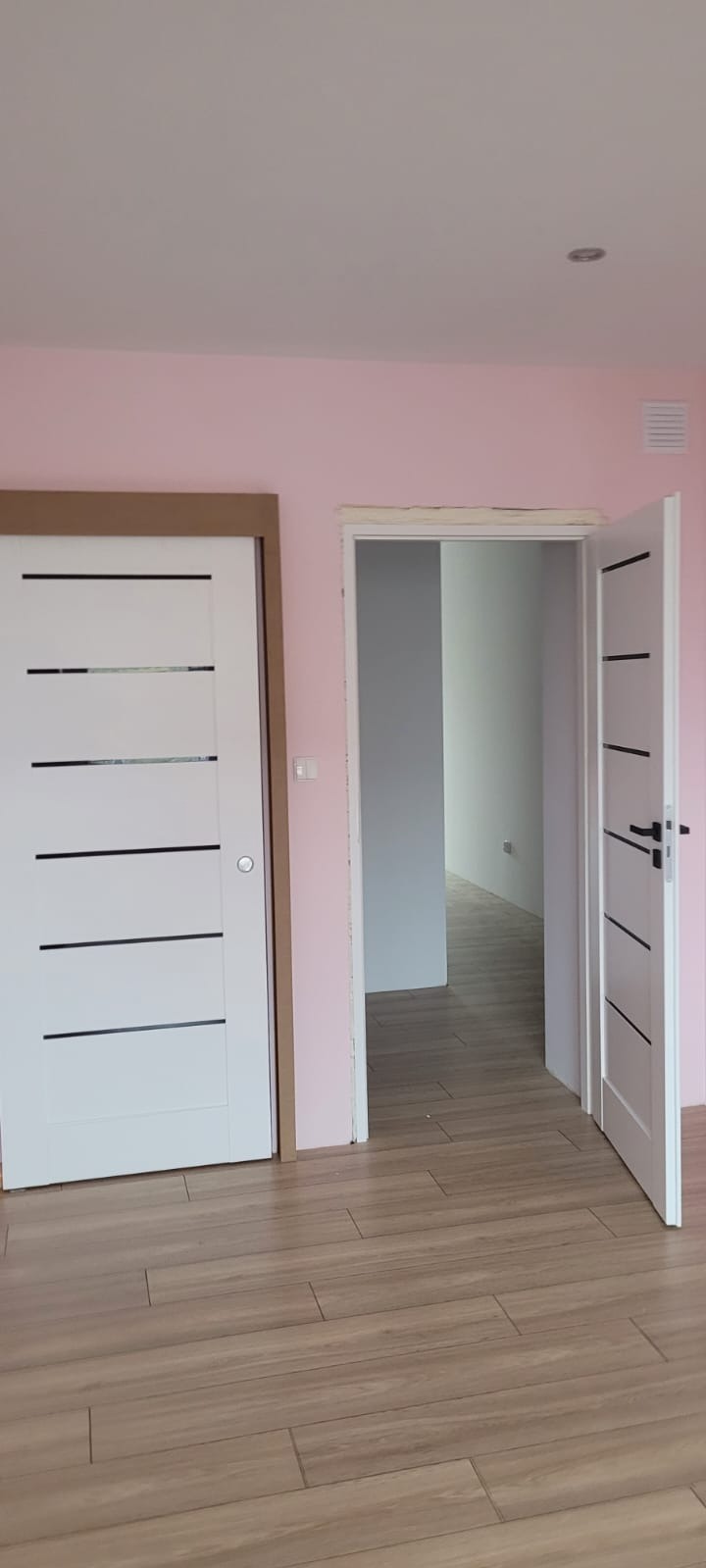 Dwa białe, nowoczesne drzwi z czarnymi akcentami na tle różowej ściany i podłogi imitującej drewno. Jedne drzwi przesuwne, drugie uchylne, otwarte.