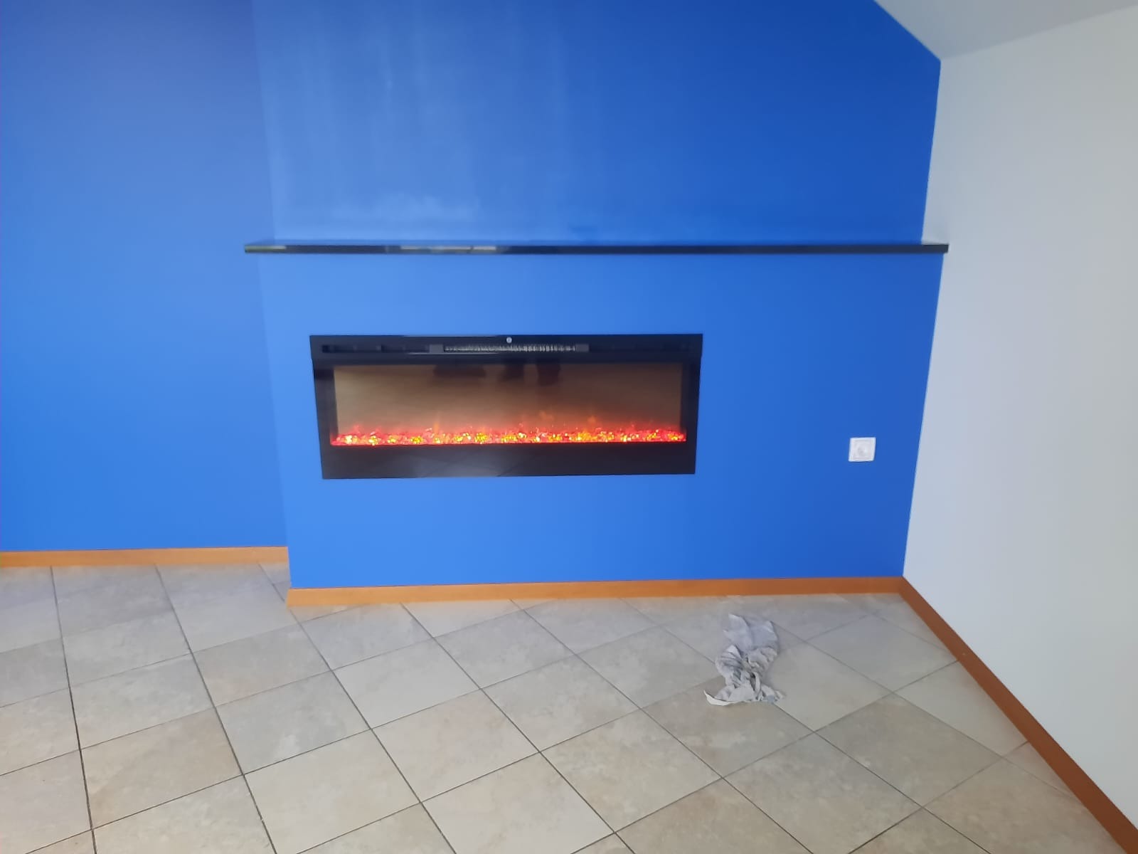 Niebieska ściana z elektrycznym kominkiem i czarną półką. Beżowa podłoga z płytek. Ścierka na podłodze. Kontrastowe połączenie kolorów ścian.