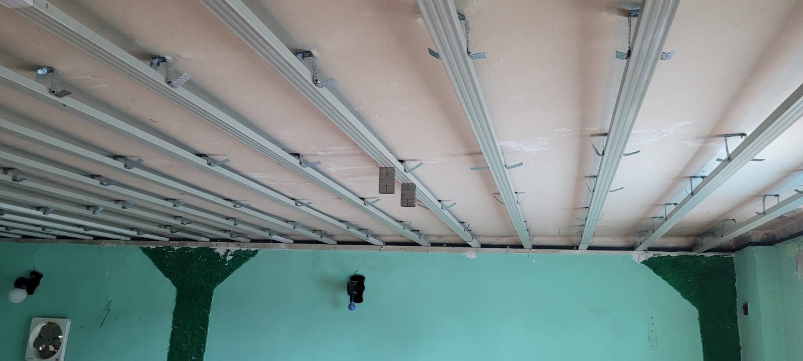 Stelaż sufitu podwieszanego z płyt karton-gips, widoczne profile i uchwyty montażowe, ściany w kolorze miętowym z dekoracyjnymi elementami.