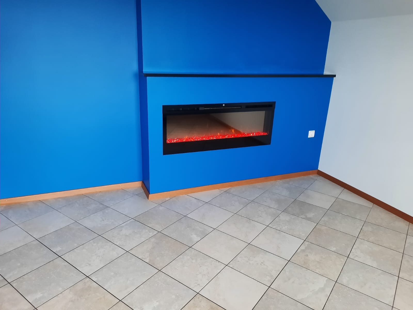 Niebieska ściana z wbudowanym elektrycznym kominkiem i beżową podłogą z płytek. Nowoczesne wykończenie wnętrza z kontrastującymi kolorami.