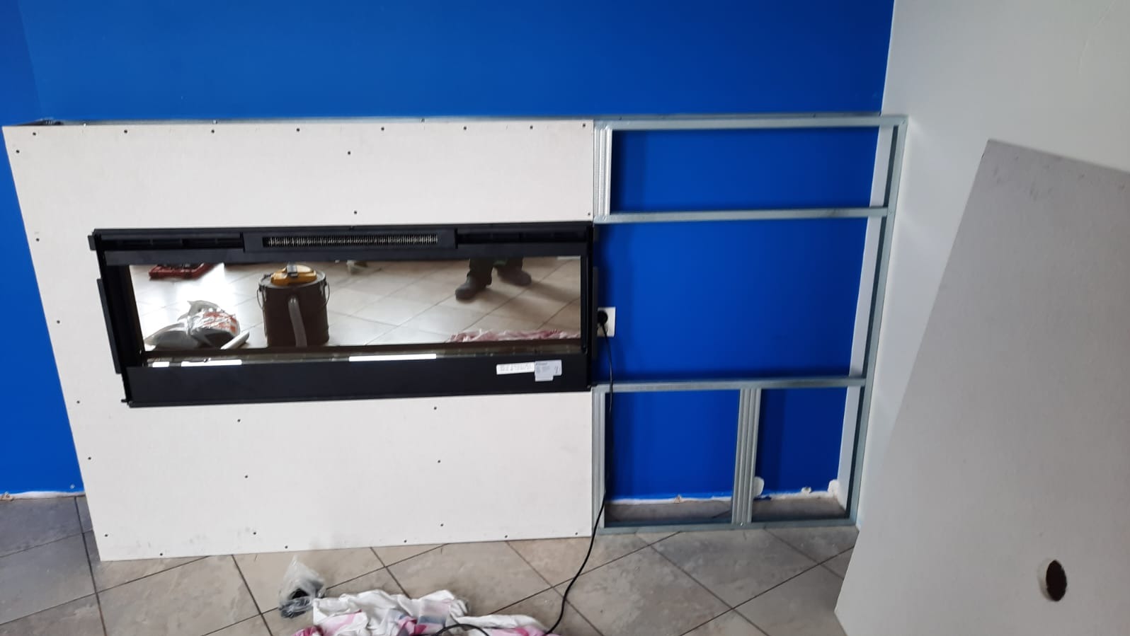 Montaż wkładu kominkowego z obudową z płyt gipsowo-kartonowych na metalowym stelażu, widoczne niebieskie ściany i fragment podłogi z płytek.