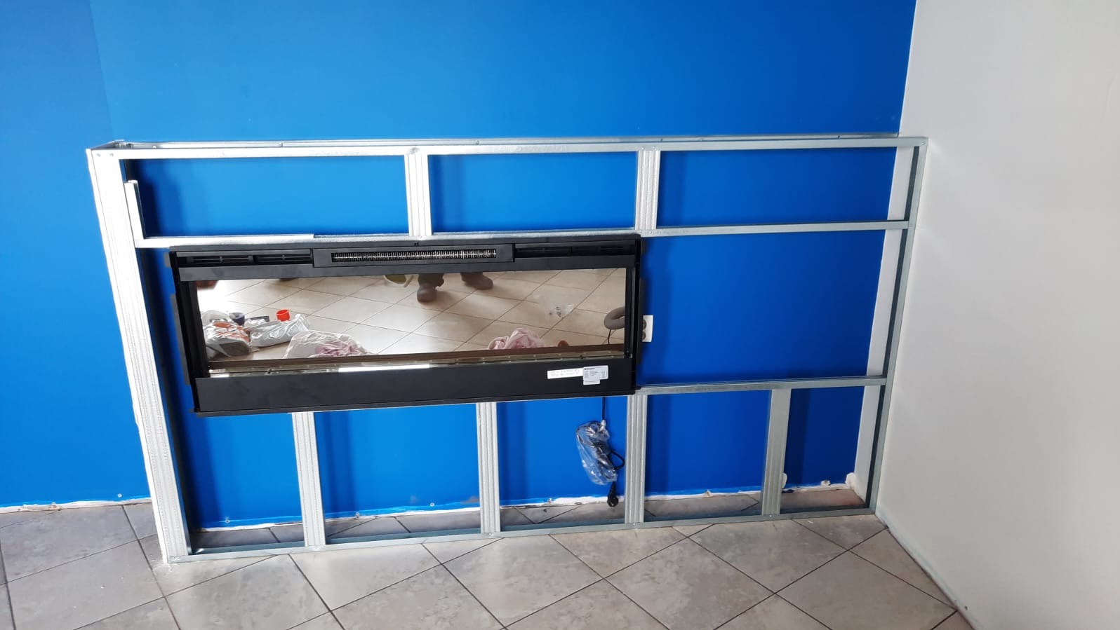 Metalowa konstrukcja pod zabudowę kominka na tle niebieskiej ściany. Widoczne profile i fragment kominka. Prace wykończeniowe w trakcie realizacji.