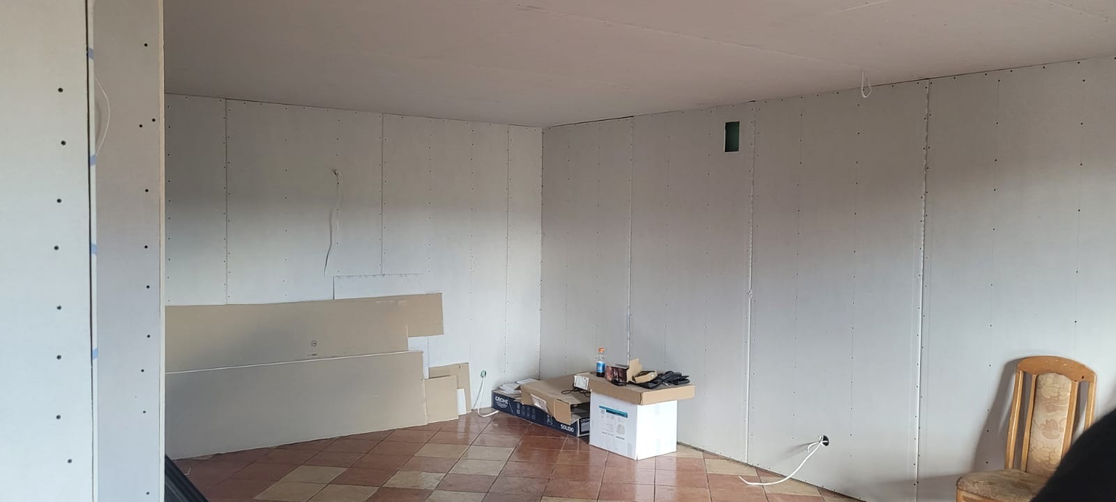 Pokój w trakcie montażu płyt kartonowo-gipsowych na ścianach. Widoczne płyty, narzędzia i instalacje elektryczne. Podłoga wyłożona płytkami.