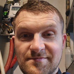 Piotr Meller Energia w Dobrym Klimacie - Mężczyzna z lampą czołową na tle zamontowanego, działającego pieca gazowego z wyświetlaczem. Widoczne przewody i elementy instalacji. Uśmiechnięty, zadowolony z wykonanej pracy.