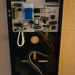 Piotr Meller Energia w Dobrym Klimacie - Otwarty panel domofonu Cyfral Smart-D z widocznymi elementami elektroniki, przewodami i numerem unifonu '11, 25' zapisanym odręcznie. Widok z bliska.