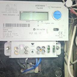 Piotr Meller Energia w Dobrym Klimacie - Szczegółowe ujęcie licznika elektrycznego ACE1000 Typ 280 z widocznymi parametrami technicznymi i podłączeniami kablowymi w skrzynce. Widoczne ślady użytkowania.