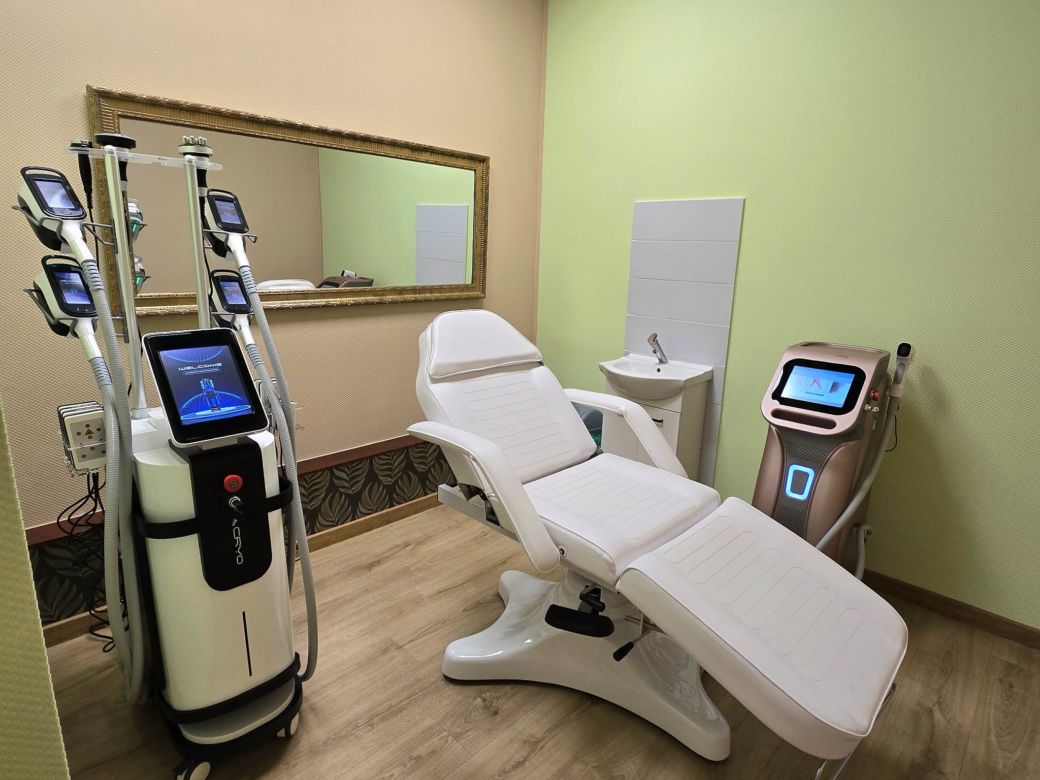 Jasny gabinet kosmetyczny z nowoczesnym sprzętem do modelowania sylwetki i depilacji laserowej. Biały fotel zabiegowy, lustro w złotej ramie, umywalka.