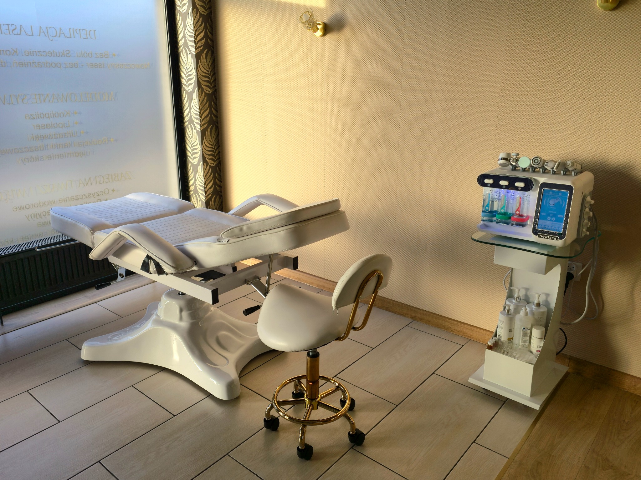 Jasne wnętrze gabinetu kosmetycznego: białe, regulowane łóżko, złoty taboret i urządzenie do zabiegów na ciało z kolorowymi roztworami. Stonowana kolorystyka.