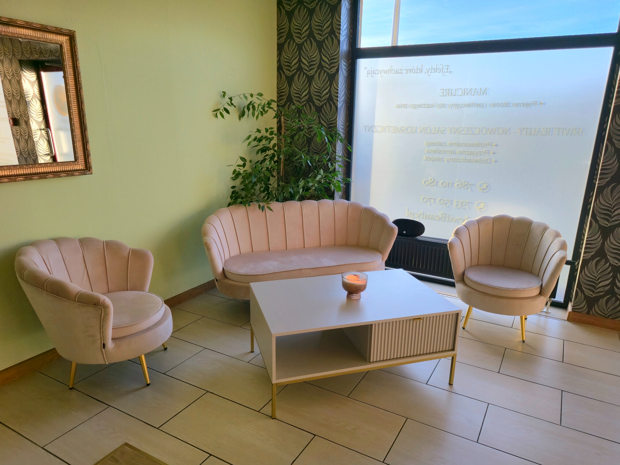 Stylowa poczekalnia salonu urody: pudrowo-różowe fotele i kanapa w stylu muszelki, biały stolik kawowy z dekoracyjną świecą, okno z reklamą salonu.