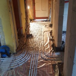 SKYBUD - Białe rury ogrzewania podłogowego ułożone na folii z czerwoną siatką w korytarzu w trakcie remontu. Widoczne ściany w stanie surowym i żółta ściana z instalacją.