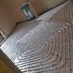 SKYBUD - Białe rury ogrzewania podłogowego ułożone spiralnie na folii z czerwoną siatką, widoczne okno w tle i fragment ściany. Etap instalacji przed wylaniem posadzki.