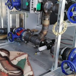 SKYBUD - Węzeł cieplny z zaworami i pompą obiegową w metalowej obudowie. Widoczne spawane rury, elementy montażowe i narzędzia. Częściowo przykryte kocem.