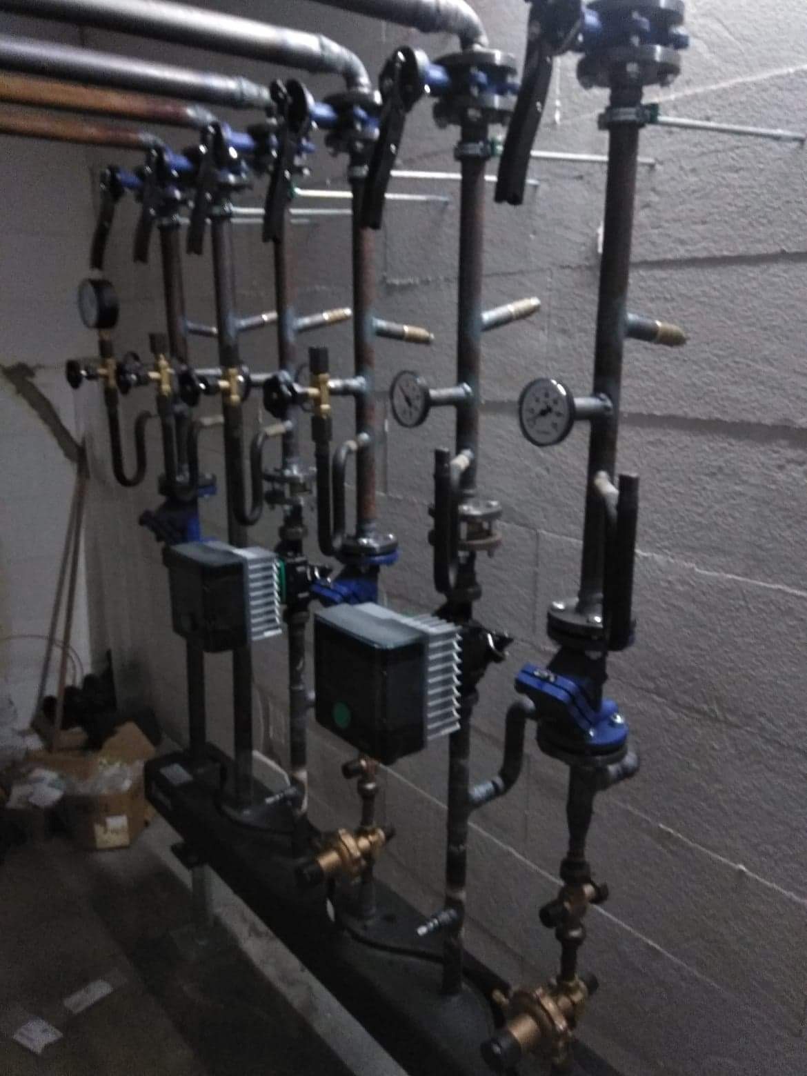 Złożona instalacja hydrauliczna z rurami, zaworami, manometrami i pompami obiegowymi, zamontowana przy szarej, teksturowanej ścianie w piwnicy lub kotłowni.