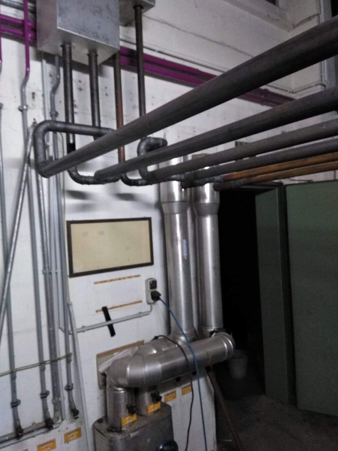 Szare rury instalacji grzewczej biegnące wzdłuż ściany z widocznym piecem i elementami podłączenia. Różne średnice rur i kształtki tworzą skomplikowany system.