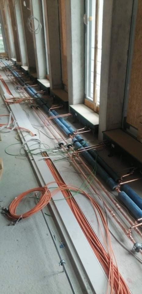 Miedziane rury instalacji grzewczej i niebieskie rury izolacyjne ułożone wzdłuż okien na etapie budowy. Widoczne przewody elektryczne i elementy konstrukcyjne budynku.