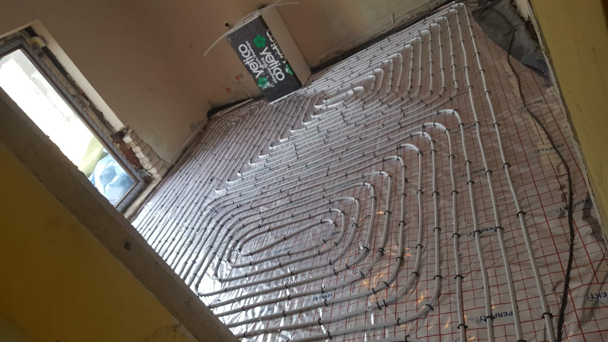 Białe rury ogrzewania podłogowego ułożone spiralnie na folii z czerwoną siatką, widoczne okno w tle i fragment ściany. Etap instalacji przed wylaniem posadzki.