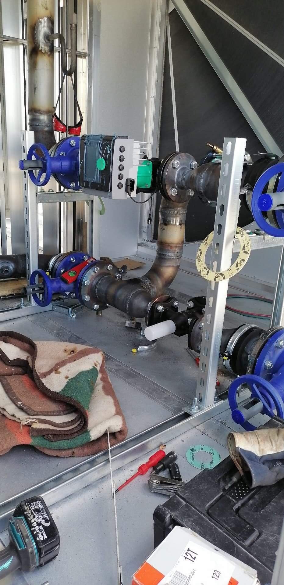 Węzeł cieplny z zaworami i pompą obiegową w metalowej obudowie. Widoczne spawane rury, elementy montażowe i narzędzia. Częściowo przykryte kocem.